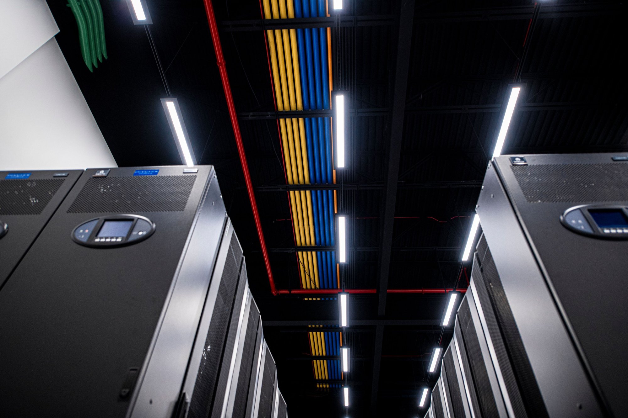 A photograph of rows of black server racks at eStruxture data center. The photograph is angled upward to show the different color-coded wiring and pipes across the ceiling in red, blue, and green, found in MTL-2 - our flagship hyperscale Montreal data center at 7001 Saint Jacques St. A photograph of rows of black server racks at eStruxture data center. The photograph is angled upward to show the different color-coded wiring and pipes across the ceiling in red, blue, and green, found in MTL-2 - our flagship hyperscale Montreal data center at 7001 Saint Jacques St.
